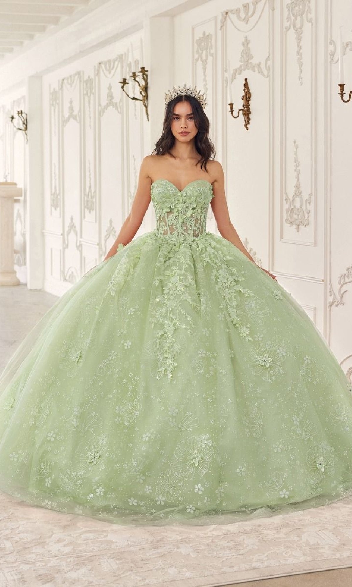 A woman in the Quinceanera Dress 15719 by Ladivine—a light green, strapless ball gown with floral embroidery and a tiara—stands in an ornate white room.